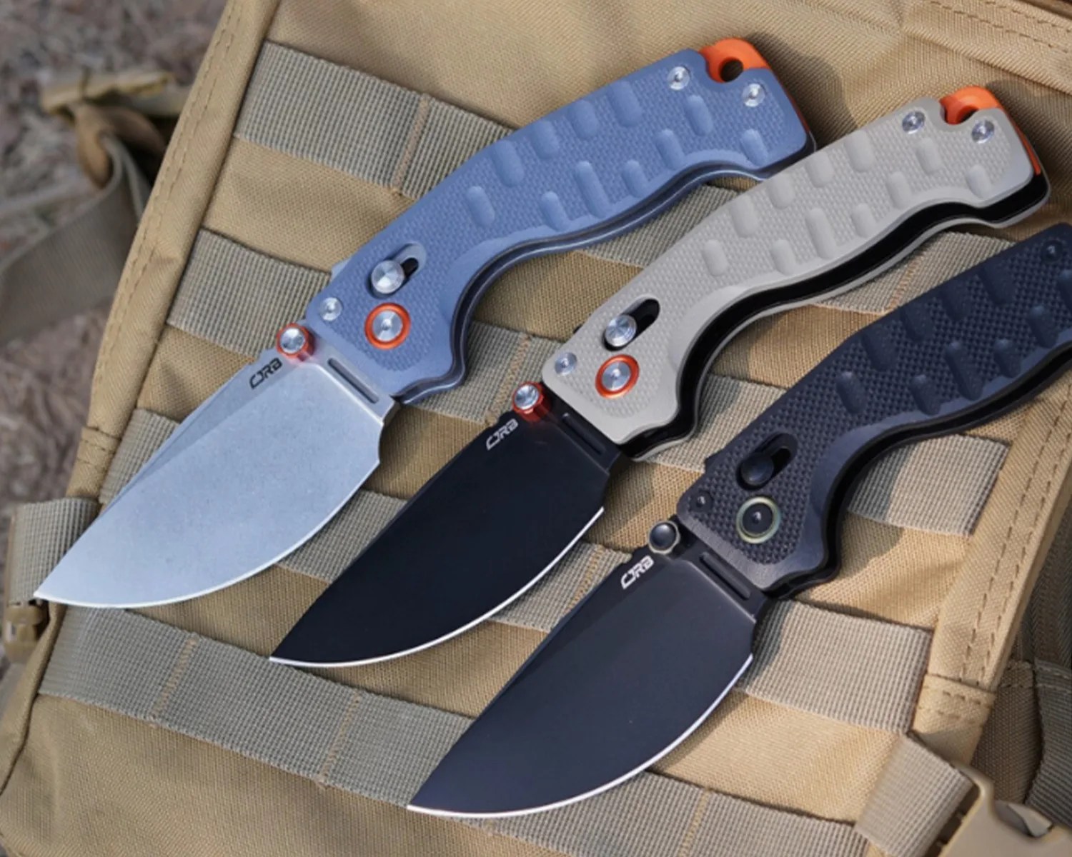 Three folding knives with textured handles in blue, beige, and black, each with a partially serrated blade, laid on a tan tactical backpack.