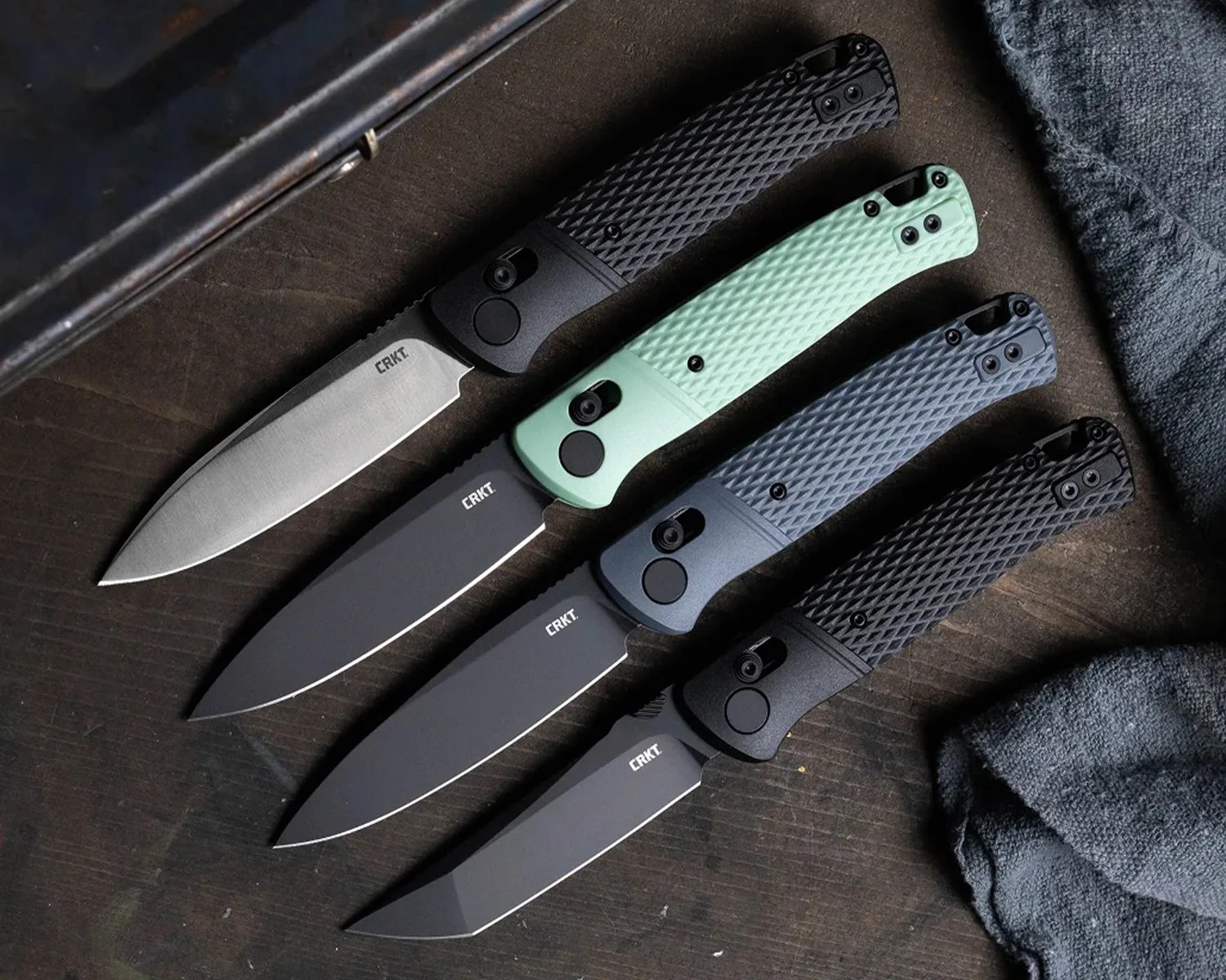 Four CRKT folding knives with textured handles in black, mint green, gray, and black, arranged on a dark wooden surface.