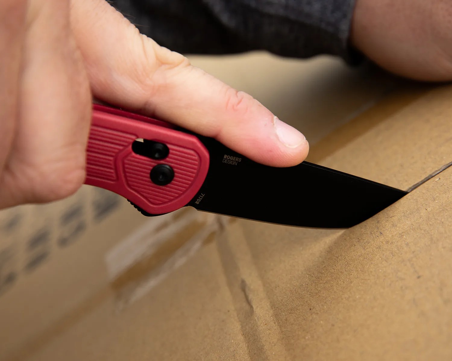 Hand holding a red-handled folding knife cutting open a cardboard box.