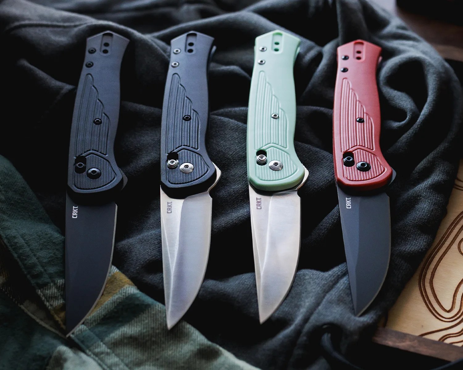 Four folding knives with textured handles in black, black, mint green, and red, laid out on dark fabric.