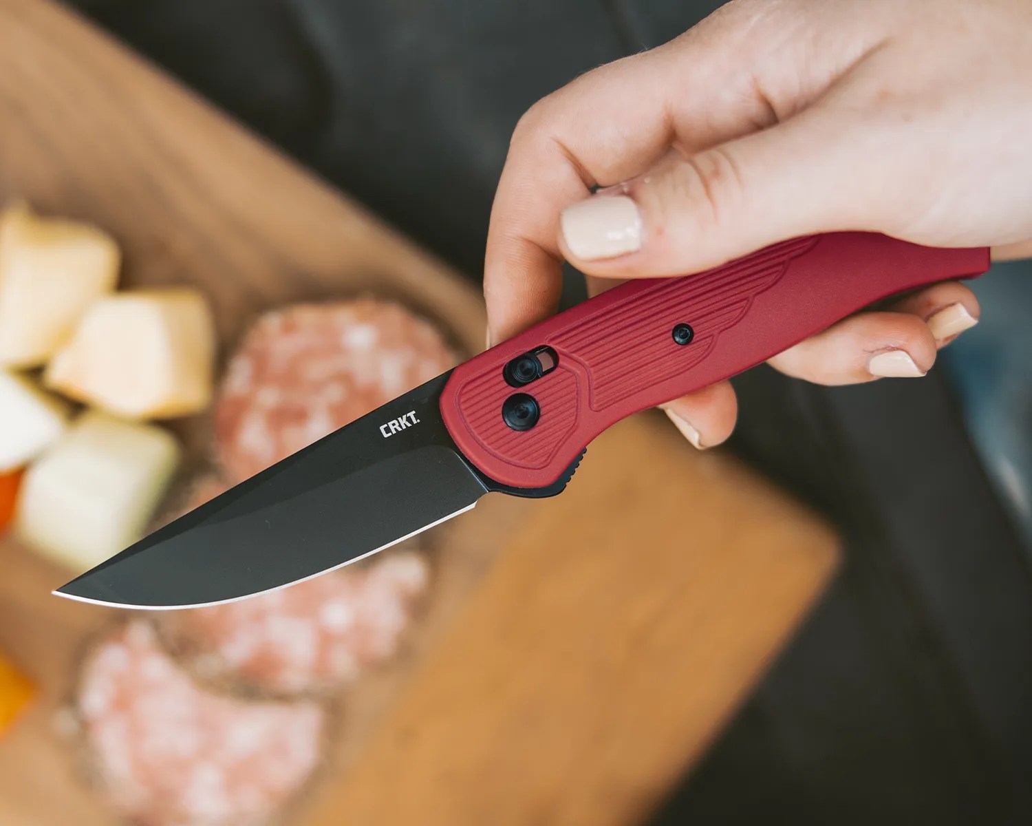 Hand holding a CRKT folding knife with a black blade and red textured handle over a wooden cutting board with sliced food.