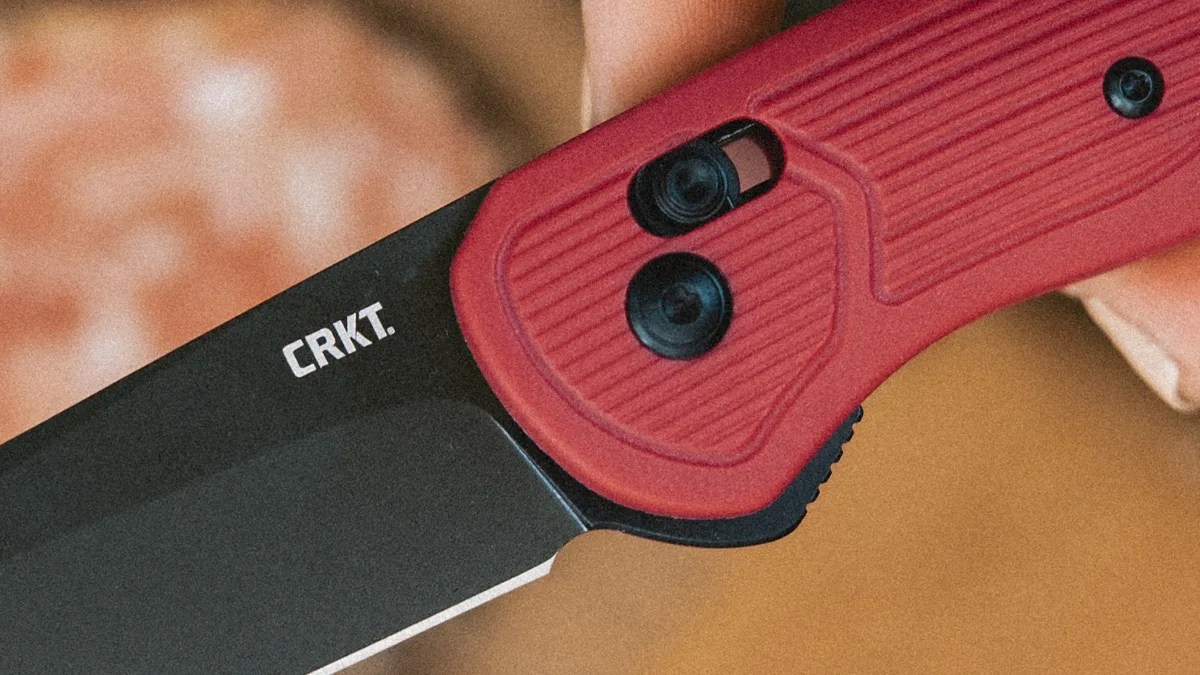 Black folding knife blade with white CRKT logo and red textured handle held in a hand.