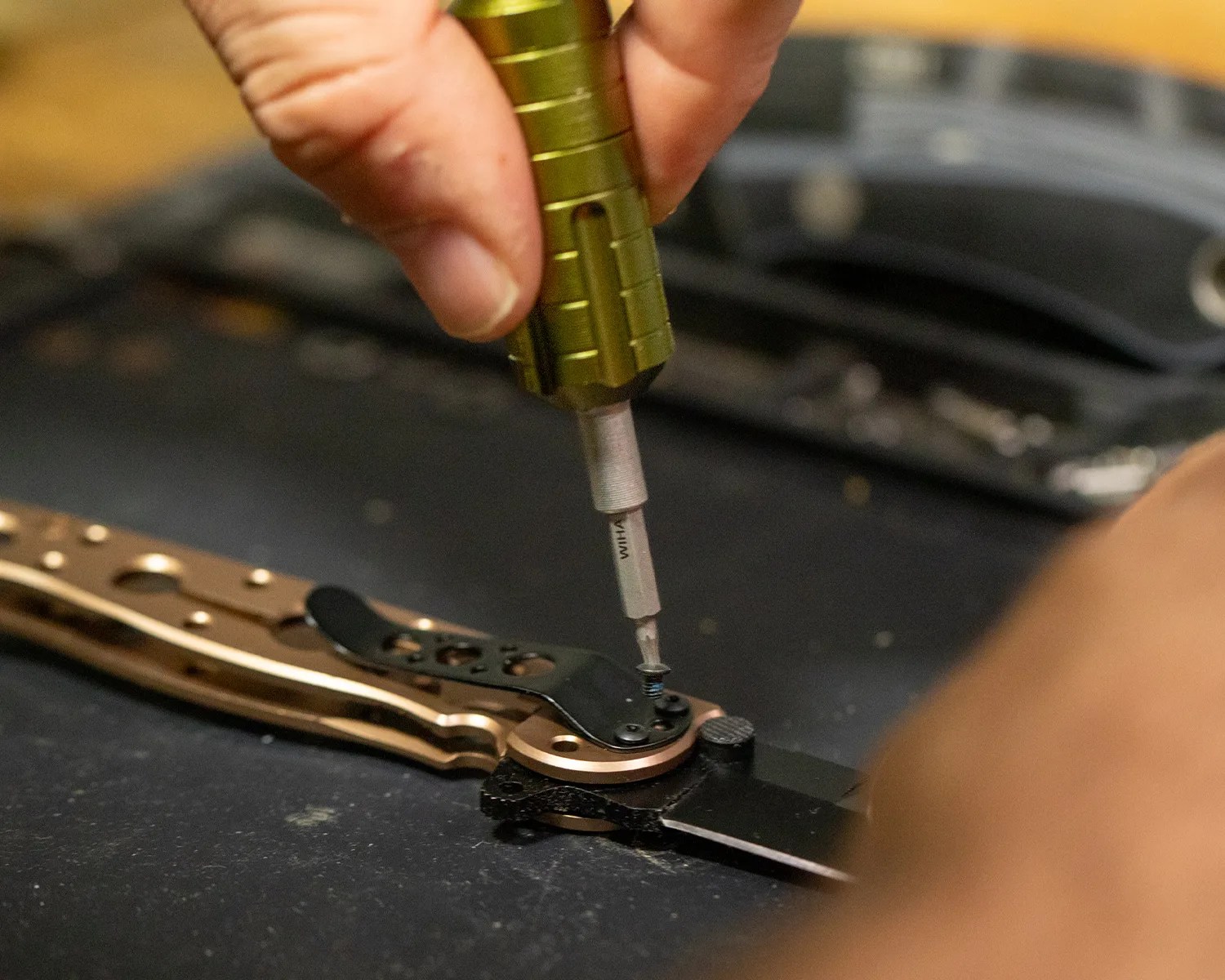 Hand using a green screwdriver to tighten a screw on a folding knife with a bronze handle.