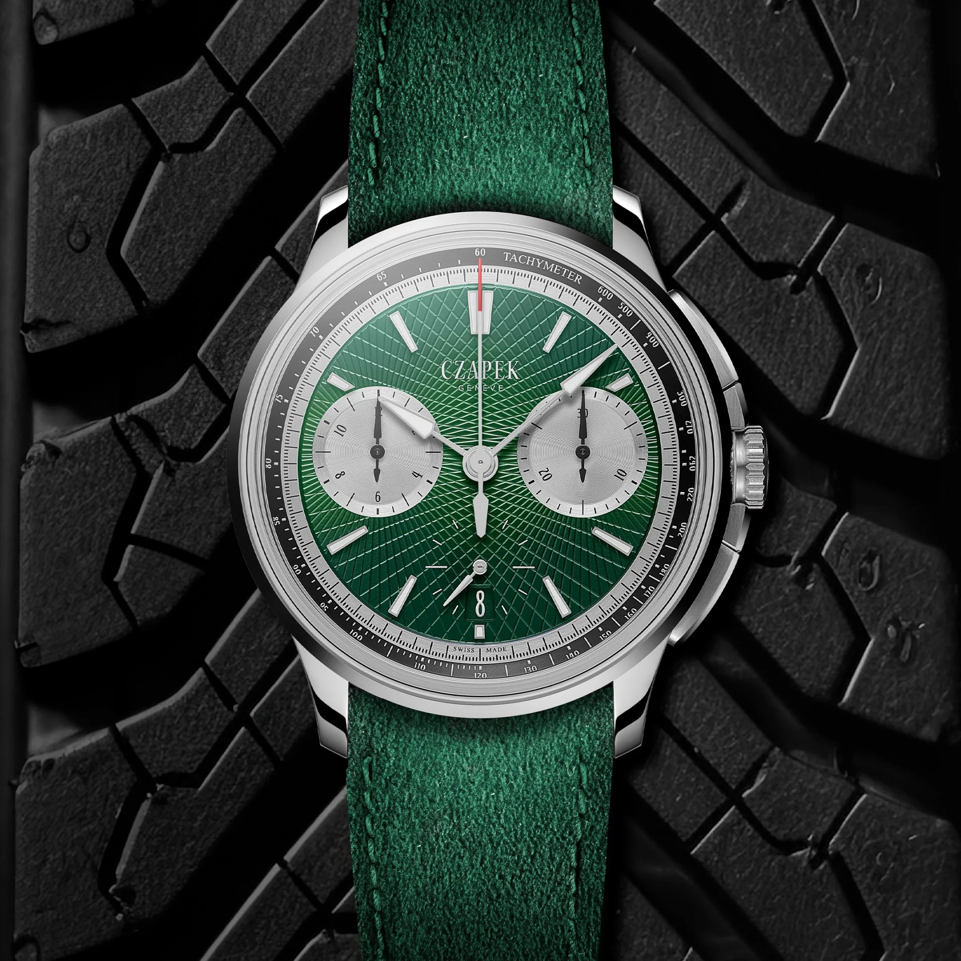 Silver chronograph wristwatch with green textured dial and matching green leather strap on black tire tread background.
