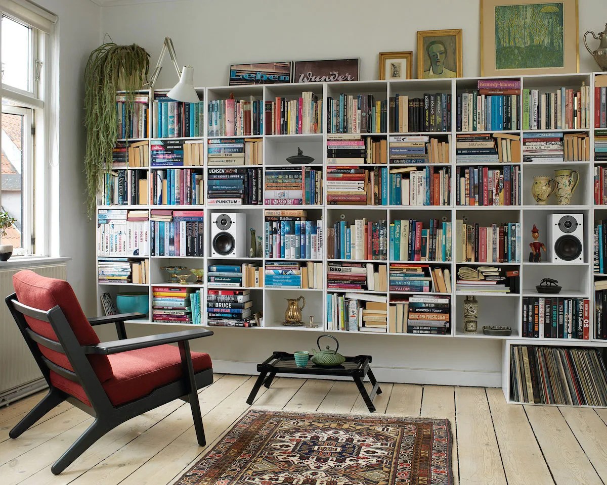 Living room with a red cushioned black wooden armchair, white wall-mounted bookshelf filled with books and decor, and a patterned rug on light wooden floor.