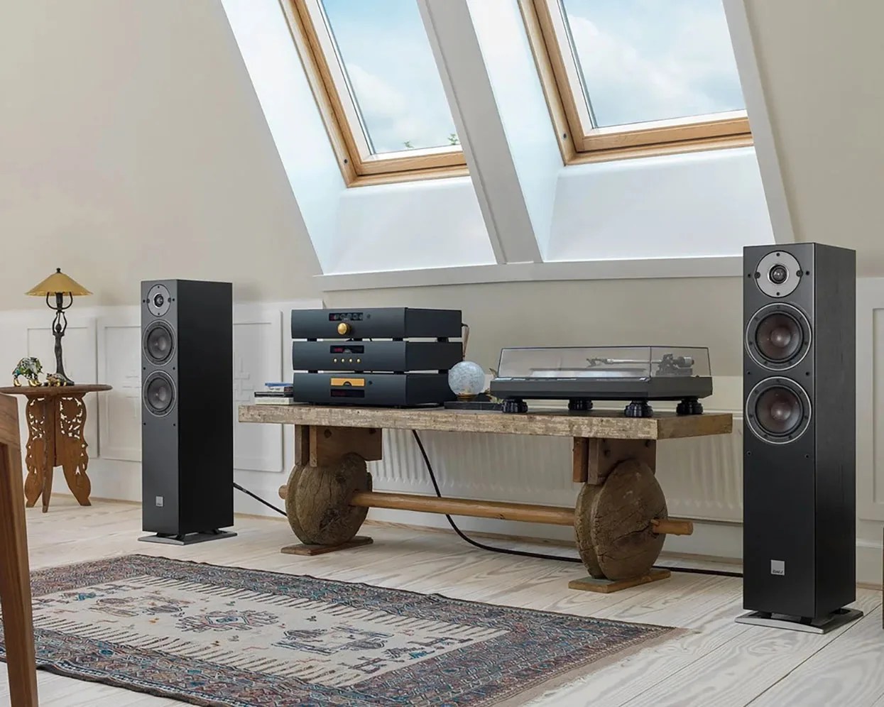 Black floor-standing speakers flanking a wooden bench with audio equipment and a turntable under skylight windows.