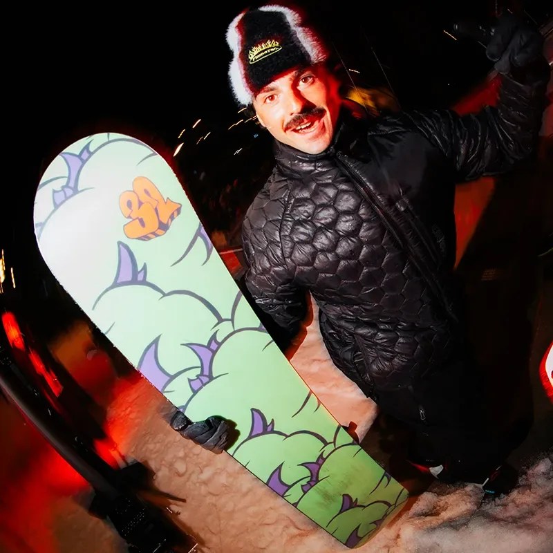 Person wearing a black quilted jacket and black gloves holding a green snowboard with purple and orange designs on snow at night.