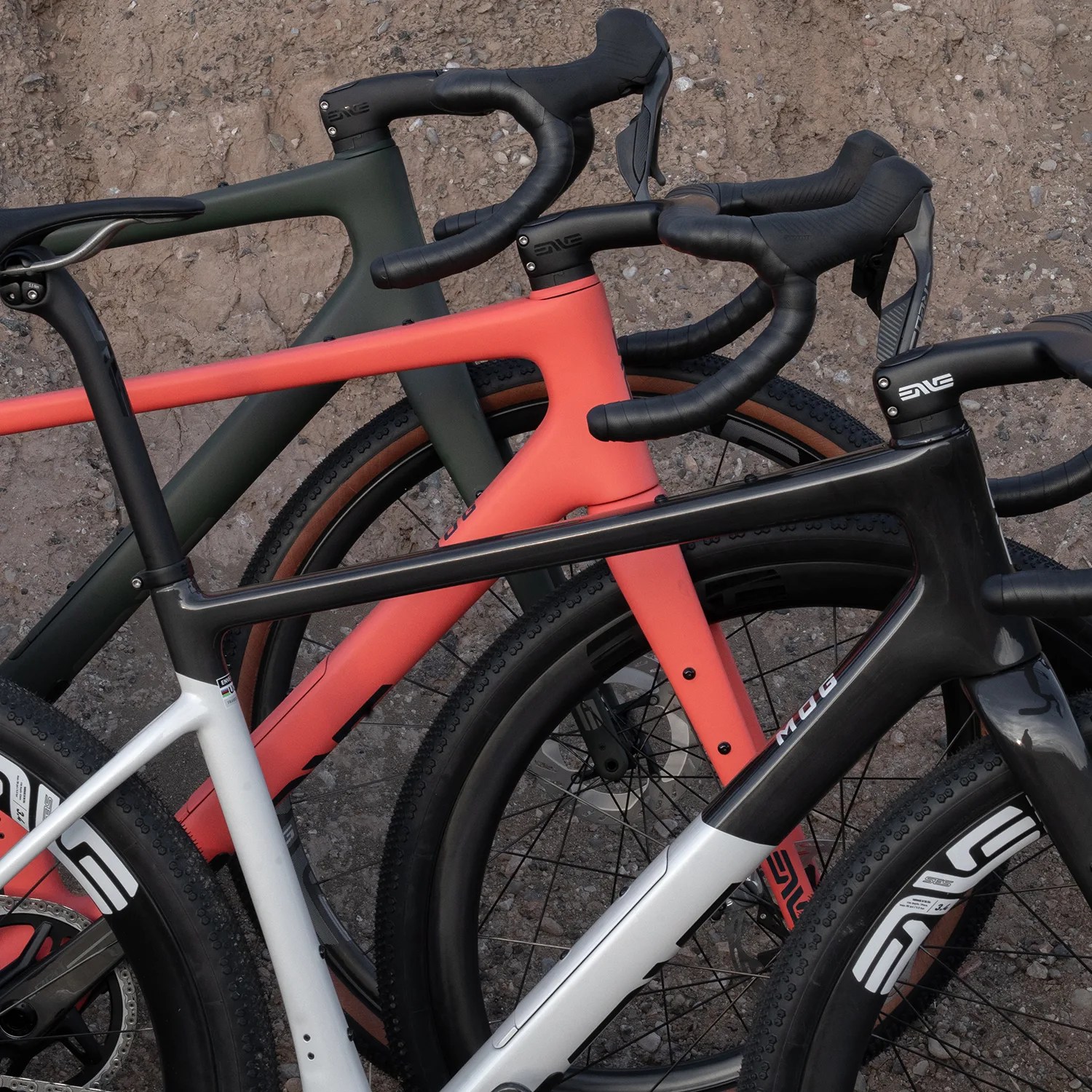 Three road bikes with drop handlebars, one black and white, one coral, and one olive green, leaning against a rough concrete wall.