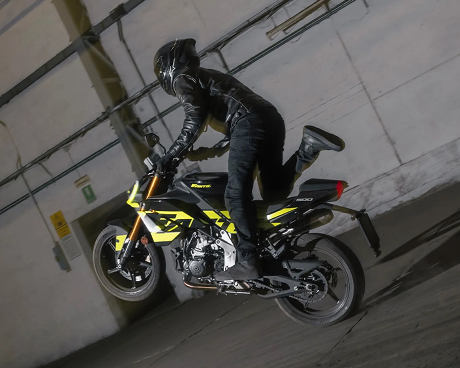 Person in black gear performing a wheelie on a black and yellow Fantic 300 motorcycle indoors.