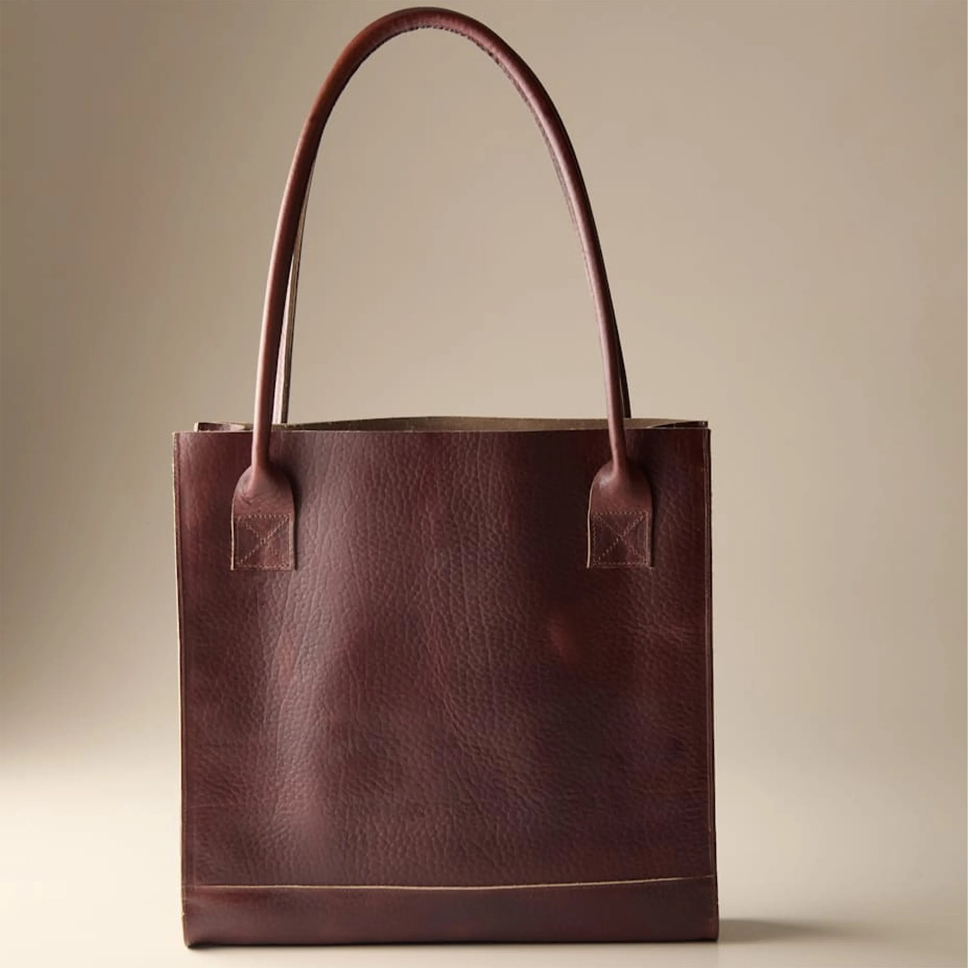 Dark brown leather tote bag with long handles against a beige background.