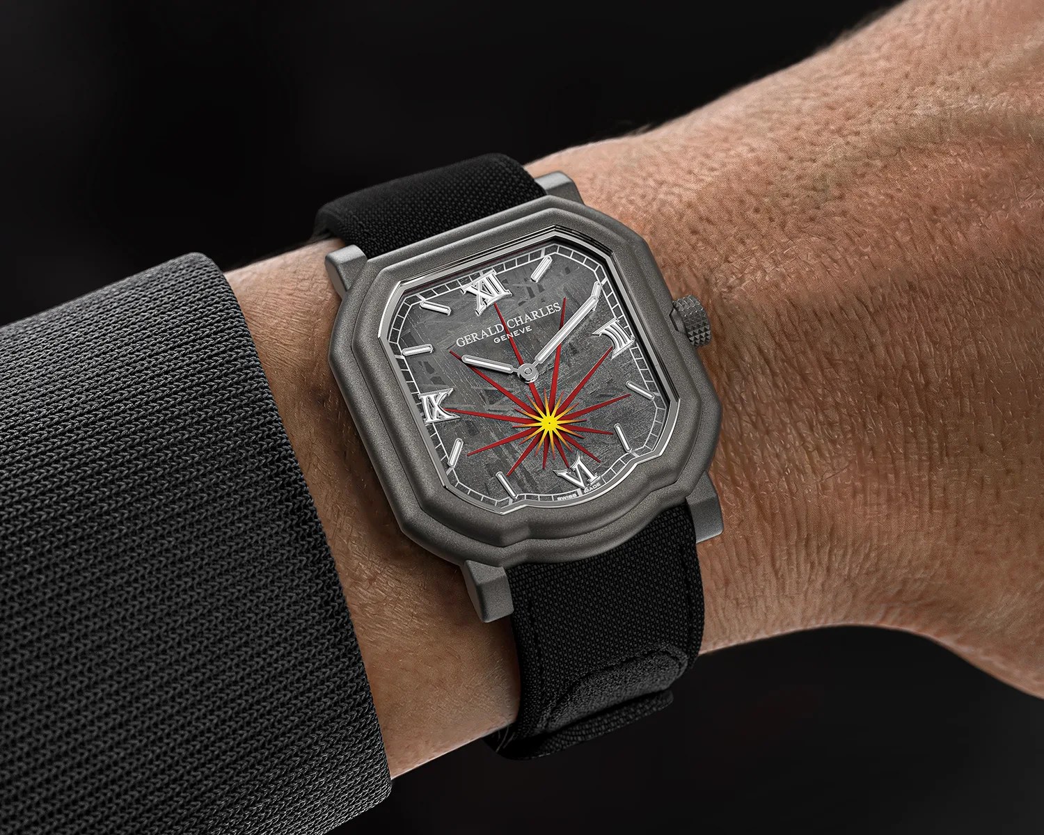 Square matte gray watch with black fabric strap and red starburst design on the dial worn on a wrist.