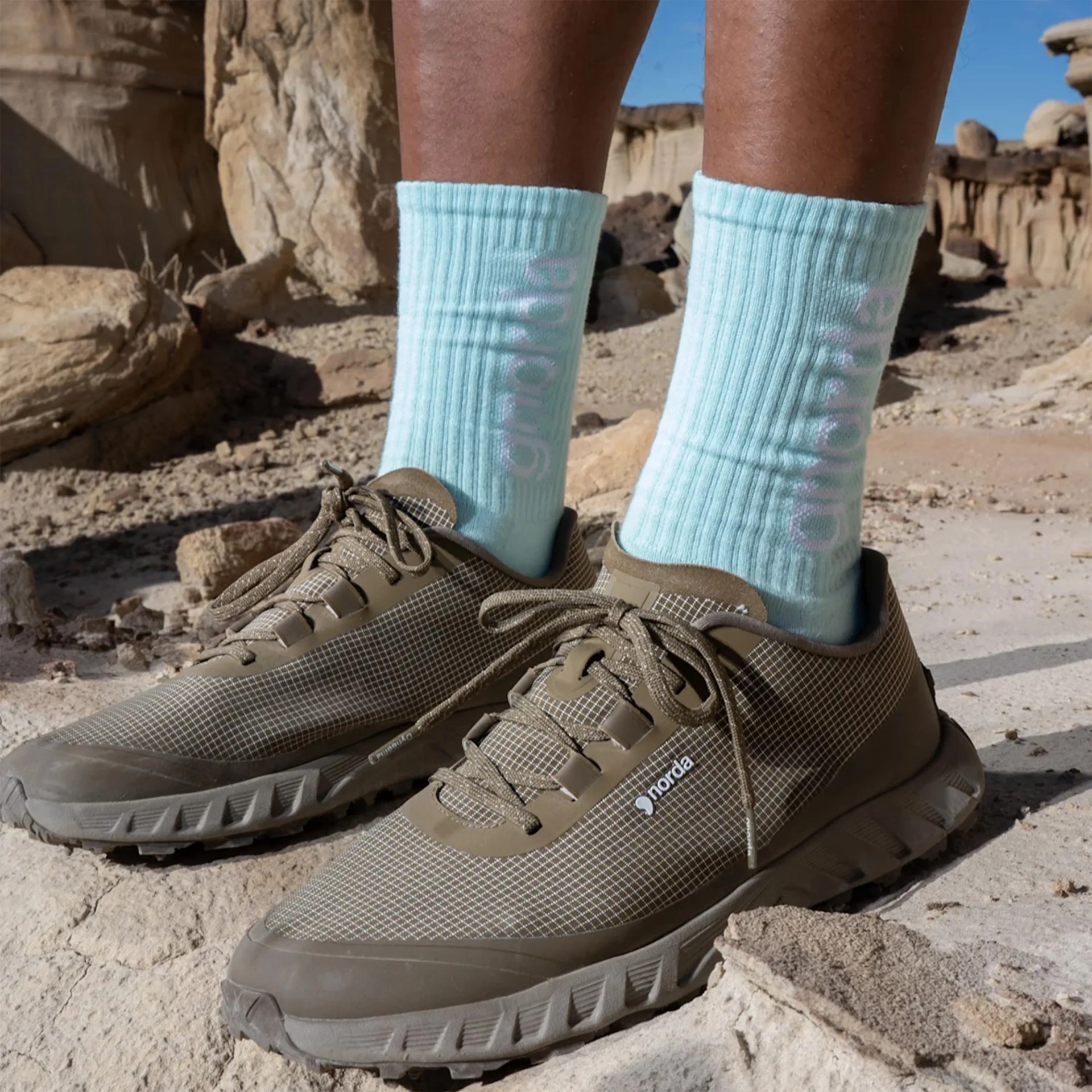 Brown Norda trail running shoes with grid pattern and laces worn with light blue Adidas socks on rocky ground.