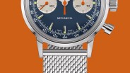 Close-up of a blue dial mechanical watch with silver mesh strap and orange chronograph hands on an orange background.
