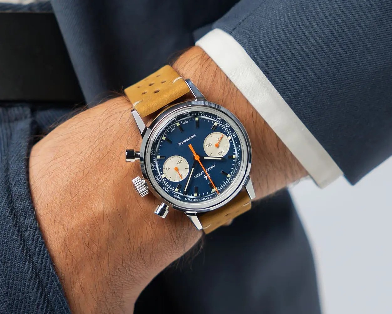 Wristwatch with blue dial, two white subdials, orange hands, and tan perforated leather strap worn with a navy suit.