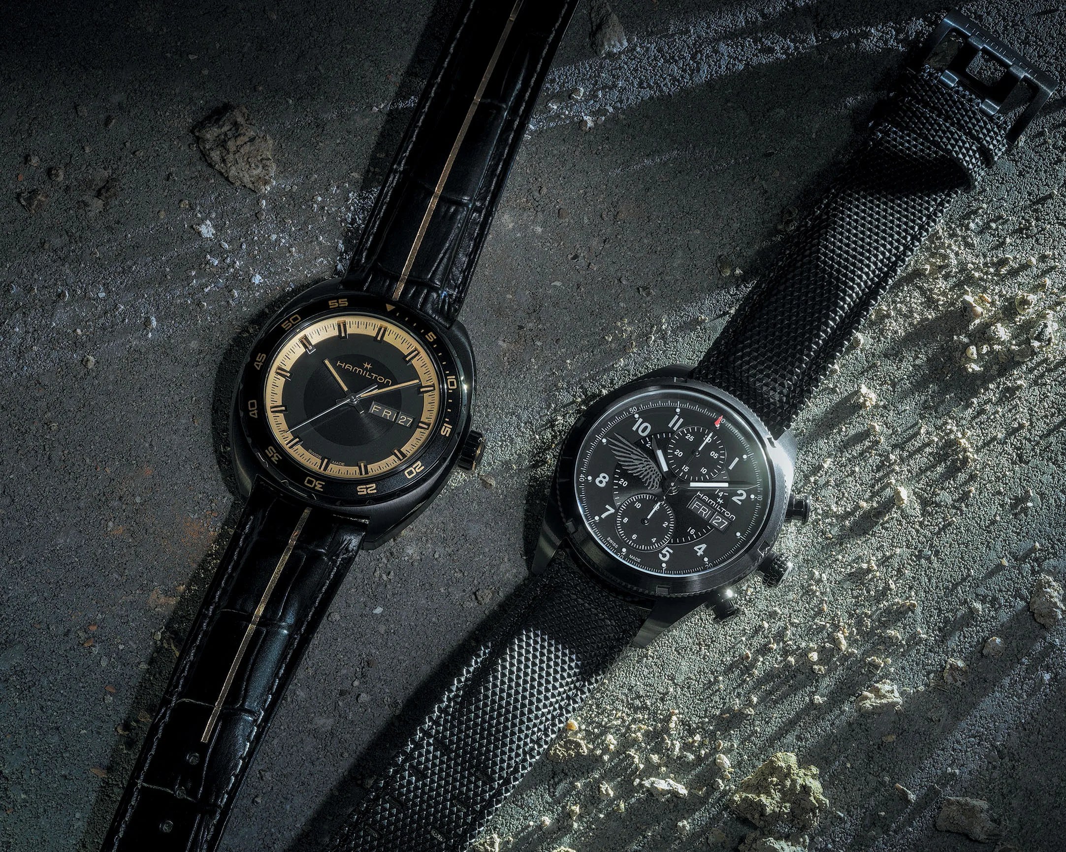Two Hamilton wristwatches on a textured concrete surface, one with a black leather strap and gold-accented dial, the other with a black textured strap and chronograph dial.