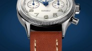 Close-up of a silver watch with a textured bezel, cream dial with blue hands, and brown leather strap.
