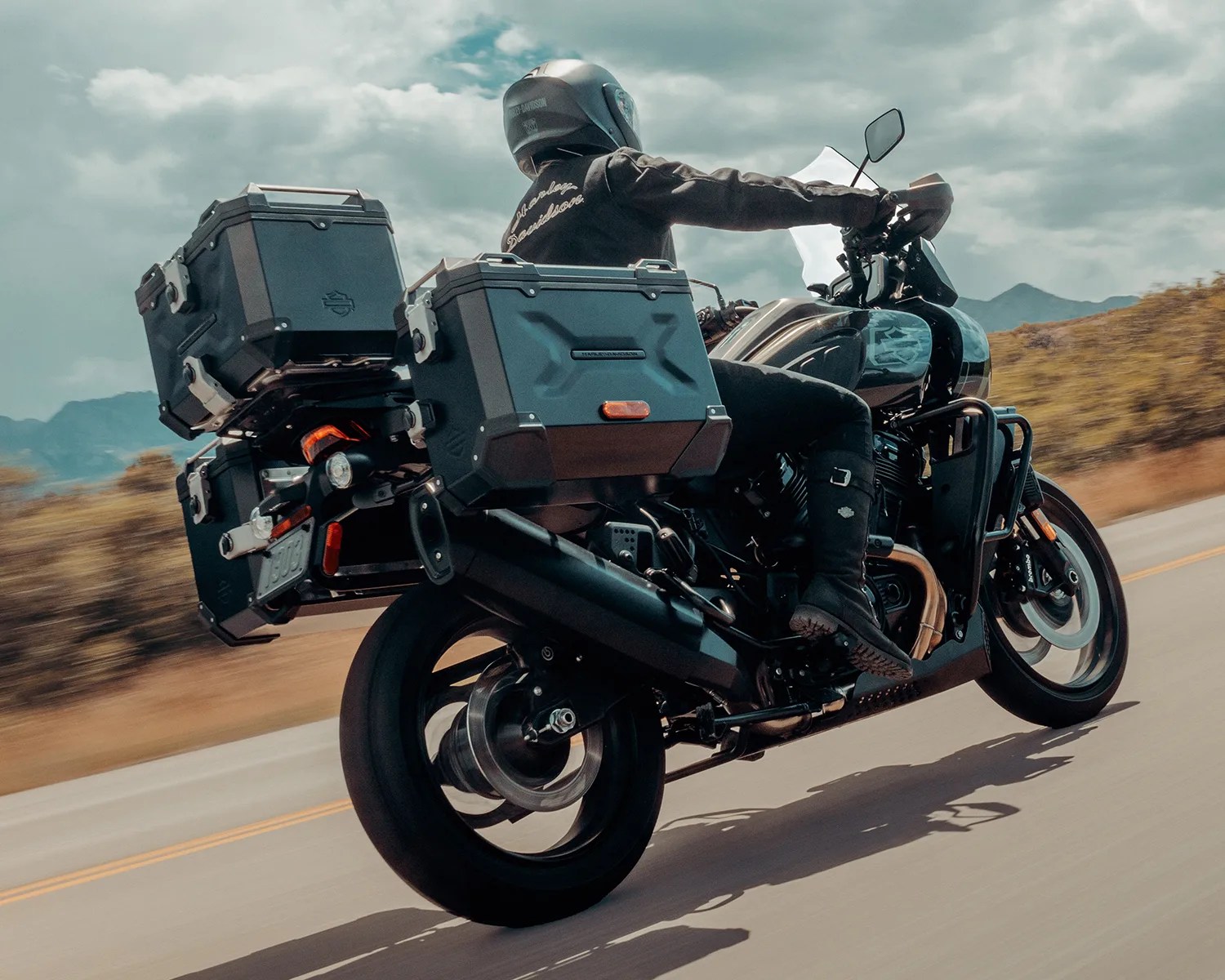 Black touring motorcycle with three large hard cases and a rider wearing a black Harley-Davidson jacket and helmet on a road.