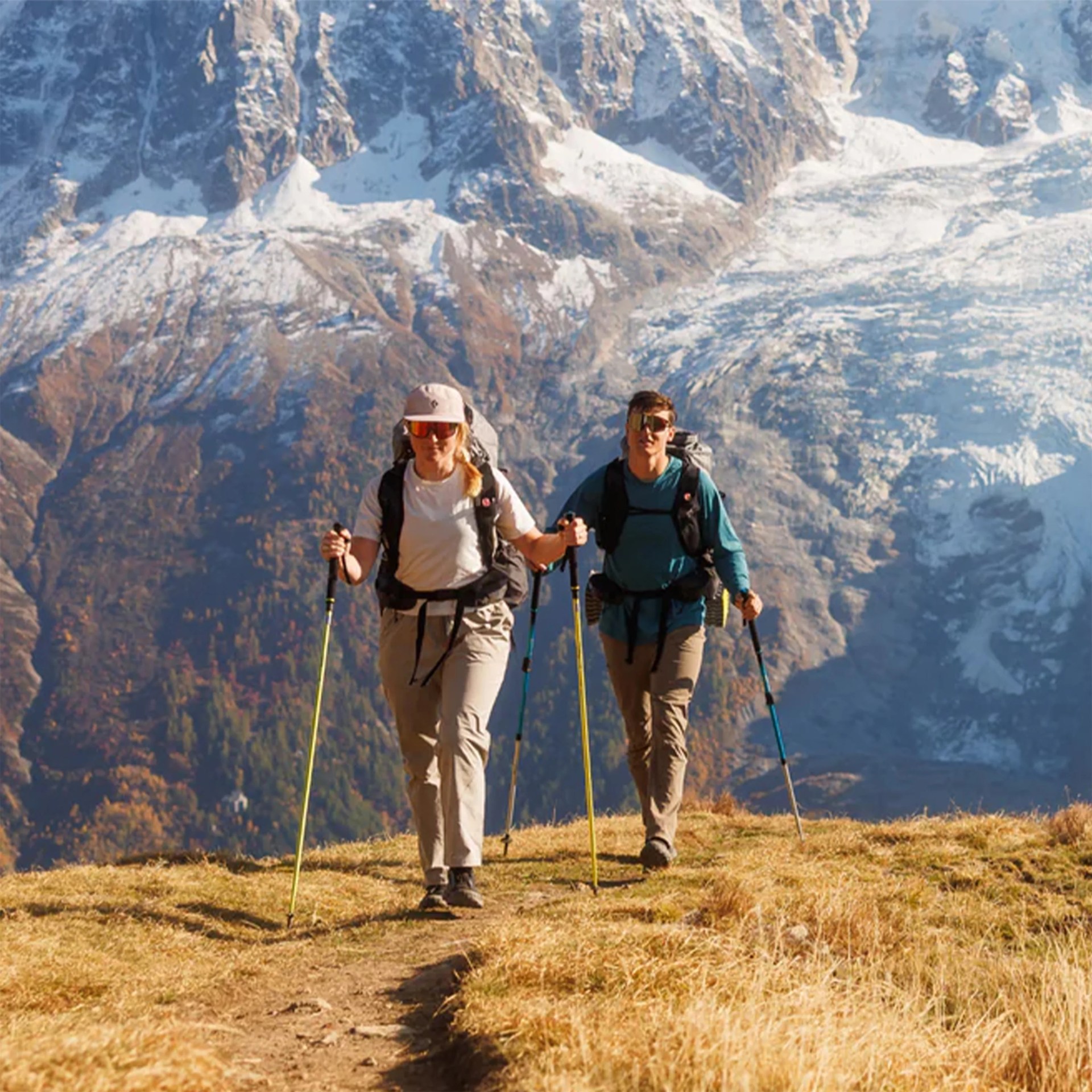 Two hikers with backpacks and trekking poles walking on a grassy mountain trail with snow-covered peaks in the background.