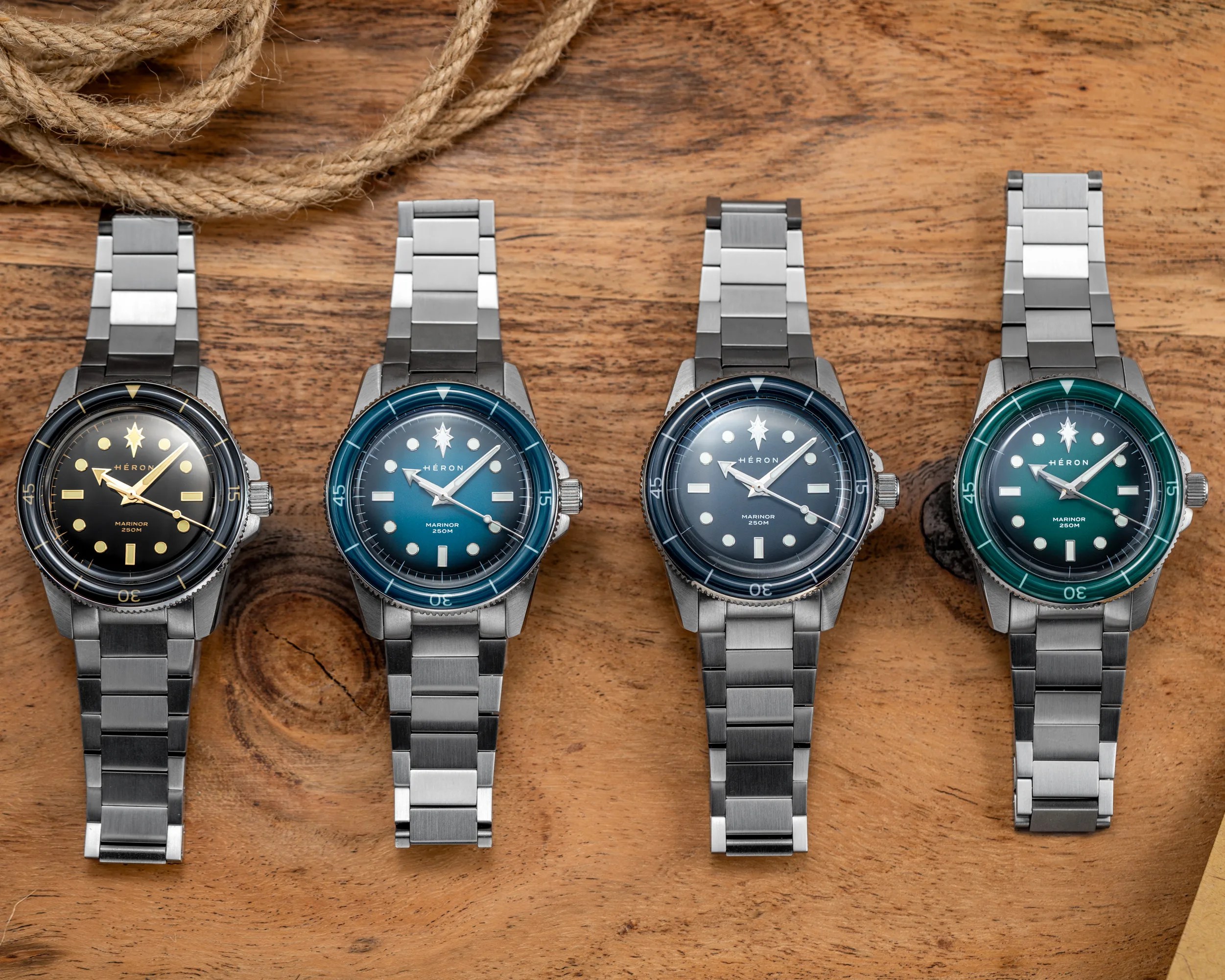 Four stainless steel wristwatches with black, blue, dark blue, and green dials on a wooden surface with coiled rope.