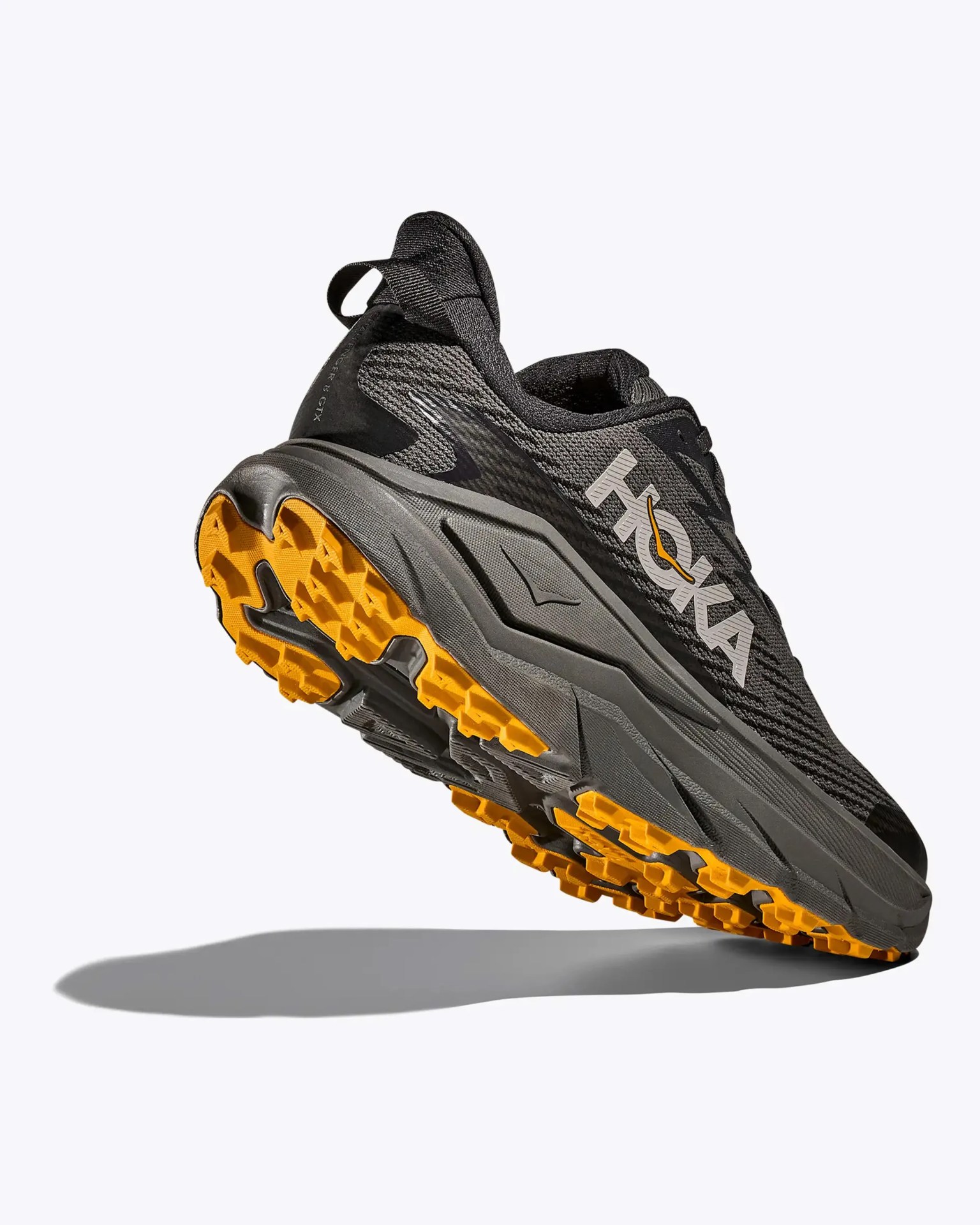 Black and gray Hoka trail running shoe with bright orange tread sole, shown from a low rear angle.