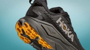 Black and gray Hoka running shoe with orange tread on the sole against a blue gradient background.