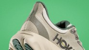 Close-up of a beige and green HOKA sneaker with textured fabric and a thick sole against a green background.