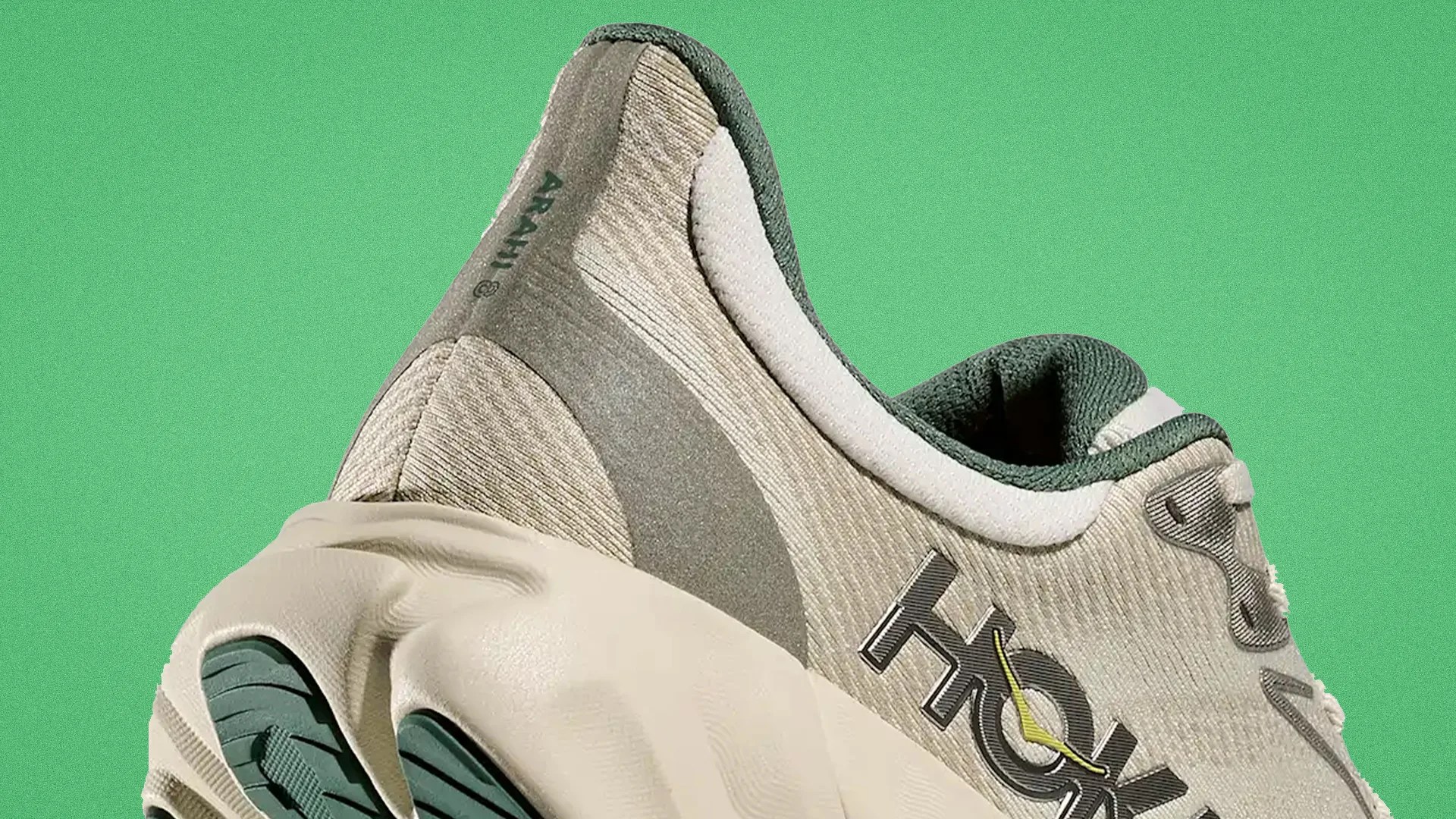 Close-up of a beige and green HOKA sneaker with textured fabric and a thick sole against a green background.