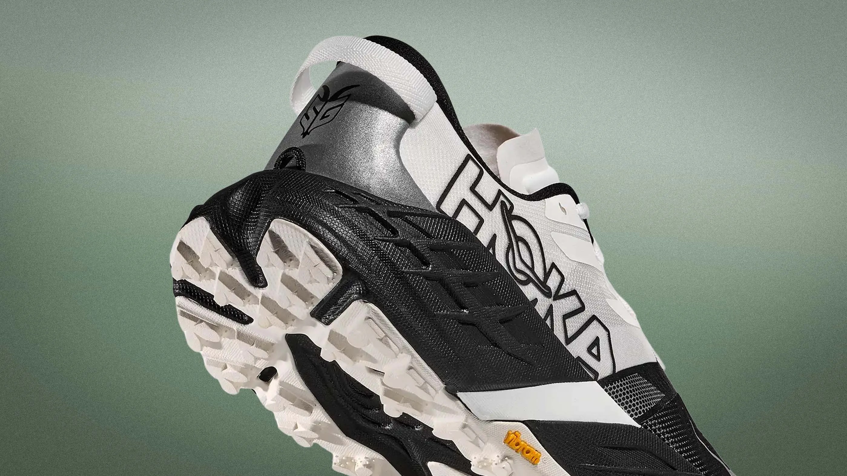 White and black Hoka running shoe with Vibram sole and silver heel tab against a green gradient background.