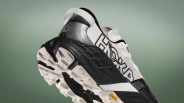White and black Hoka running shoe with Vibram sole and silver heel tab against a green gradient background.