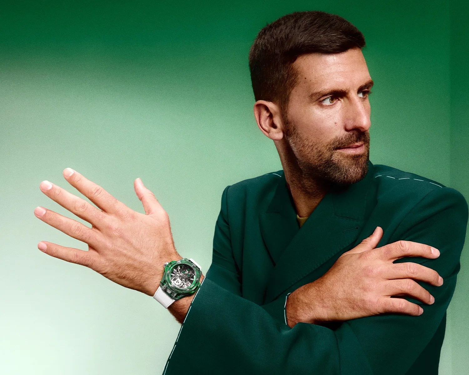 Man in a dark green blazer wearing a transparent green and white wristwatch on his left wrist against a green gradient background.