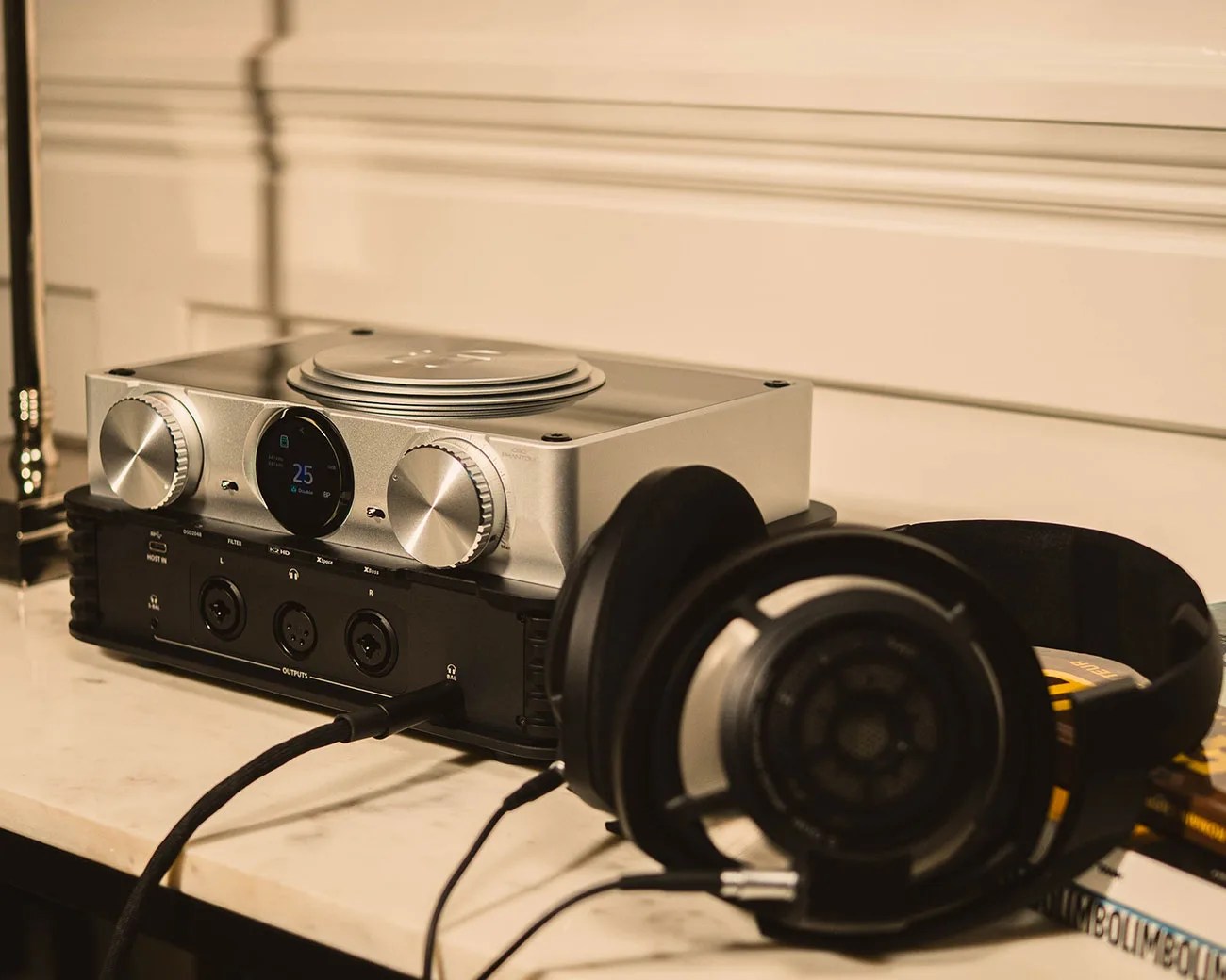 Silver audio amplifier with two large knobs and a digital display stacked on a black audio interface, with black over-ear headphones resting nearby on a marble surface.