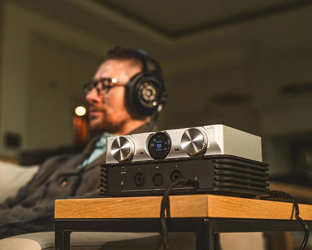 Silver and black audio amplifier with knobs and digital display on a wooden table, with a person wearing headphones in the background.