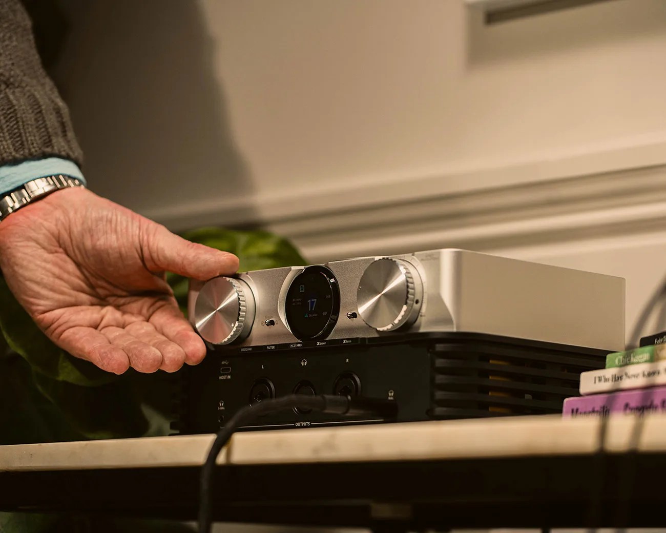 Hand adjusting the left knob on a silver and black audio amplifier with a digital display on a table.