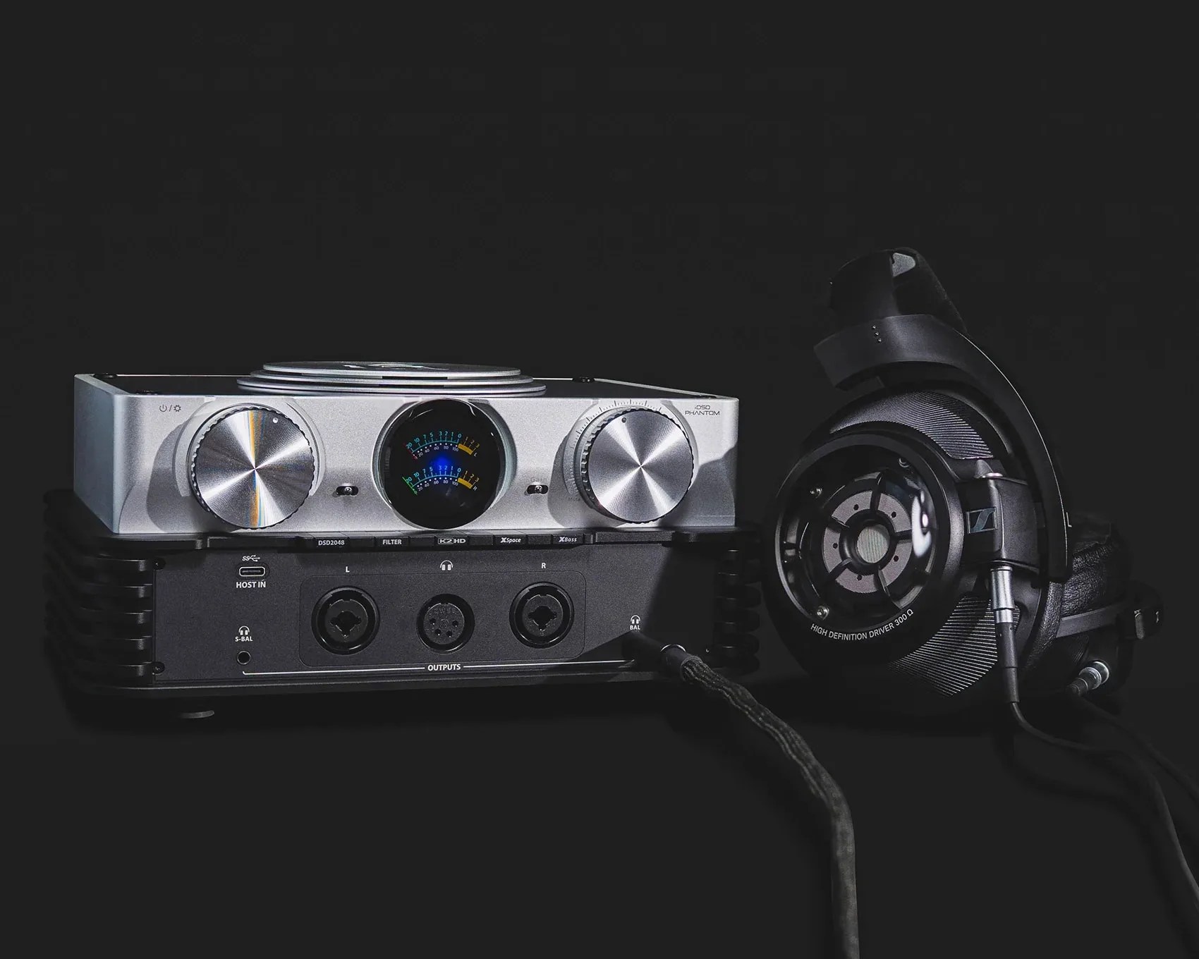 Silver audio amplifier with large knobs and analog meters next to black over-ear headphones with visible drivers.