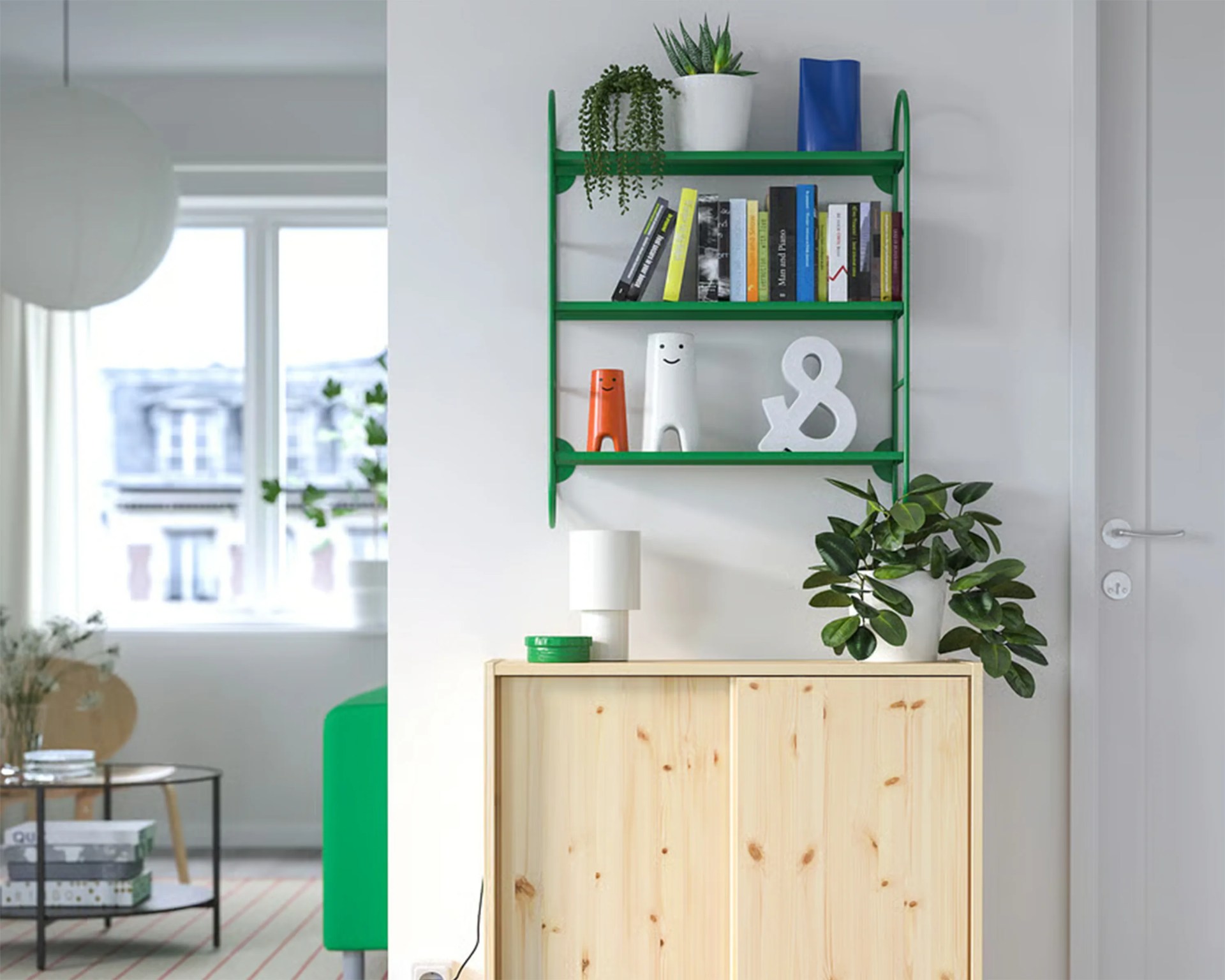 Green wall-mounted shelf with books, plants, and decorative items above a light wood cabinet with a white lamp and potted plant.