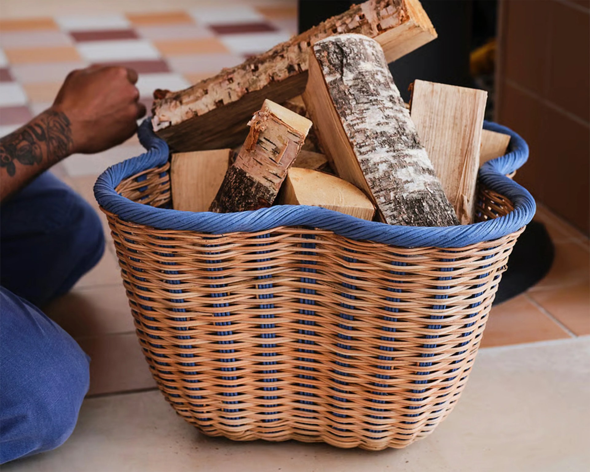 Woven wicker basket with blue trim filled with chopped firewood on a tiled floor.