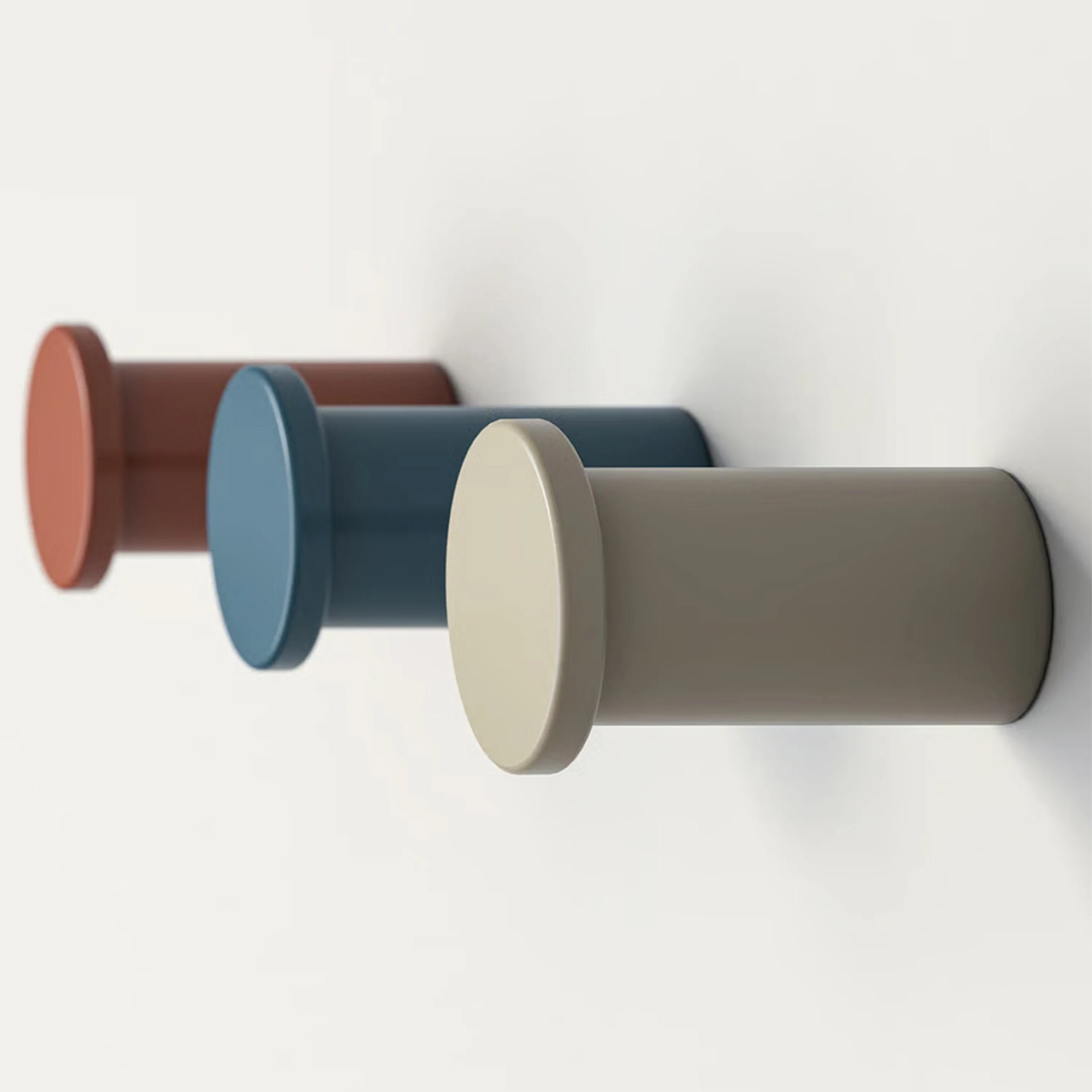 Three cylindrical wall hooks in beige, blue, and terracotta colors mounted on a white wall.