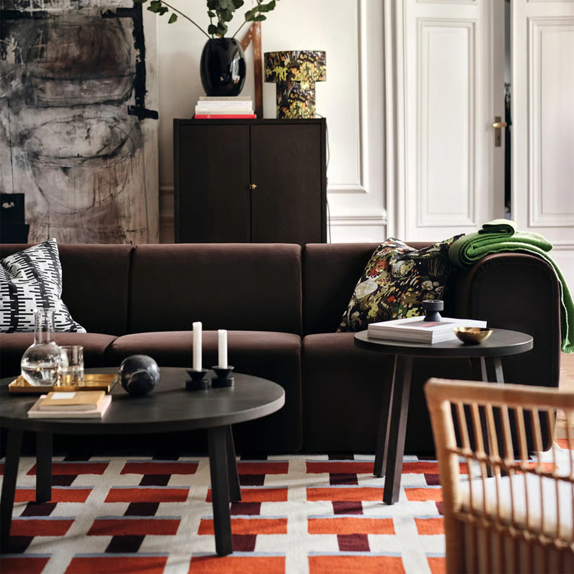 Dark brown sectional sofa with patterned pillows and green throw blanket, black round coffee tables, and geometric red and white rug.