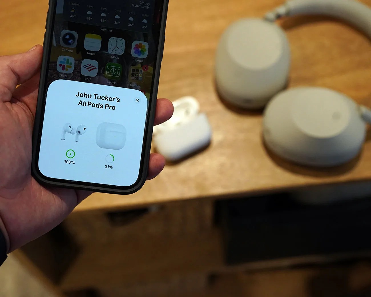 Hand holding a smartphone displaying AirPods Pro battery status with white AirPods case and beige headphones on a wooden table.