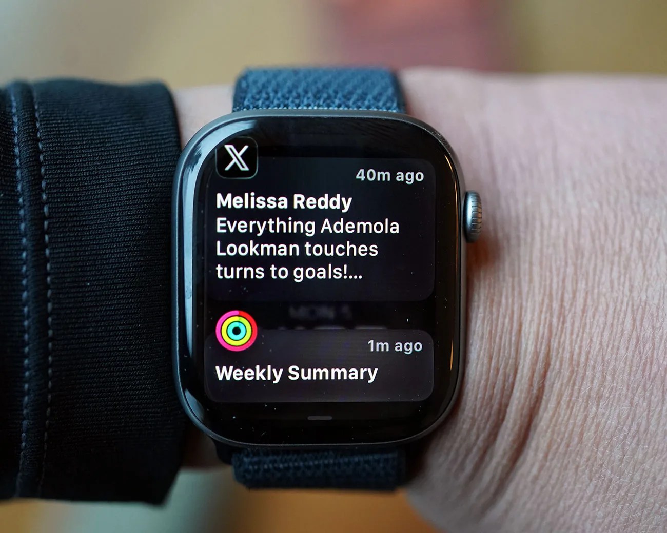 Smartwatch with a blue fabric band displaying notifications including a message from Melissa Reddy and a weekly summary.