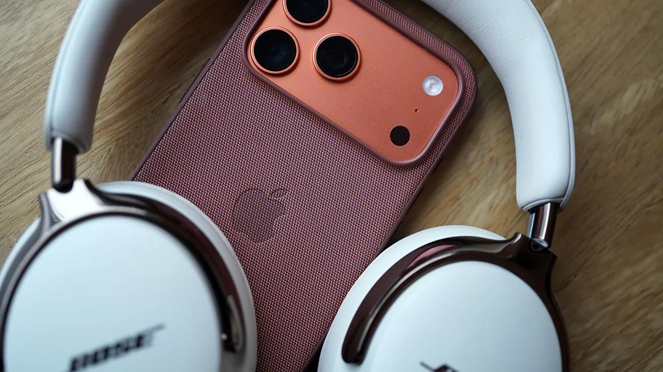 Copper-colored iPhone with a textured case lying on a wooden surface, partially surrounded by white Bose headphones.