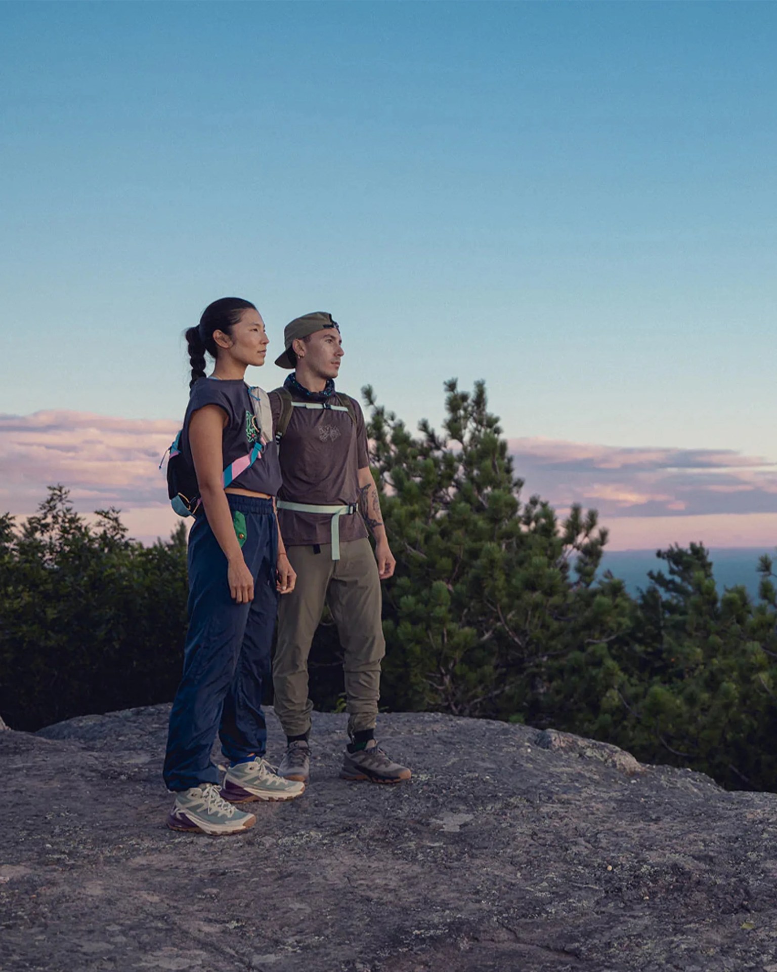 Two hikers wearing backpacks and outdoor clothing standing on a rocky surface with trees and a sunset sky in the background.