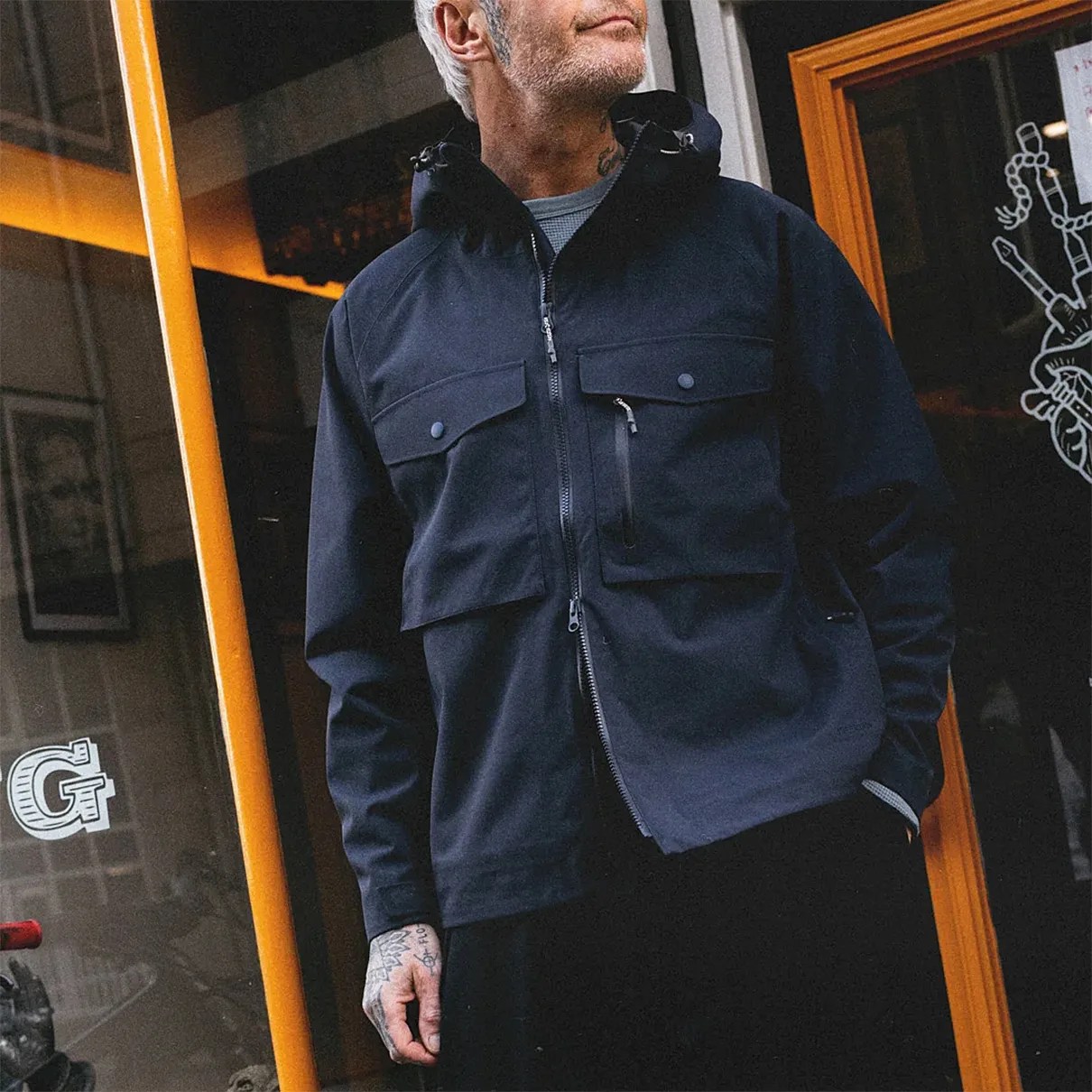 Man wearing a black hooded jacket with multiple front pockets and a zipper closure.