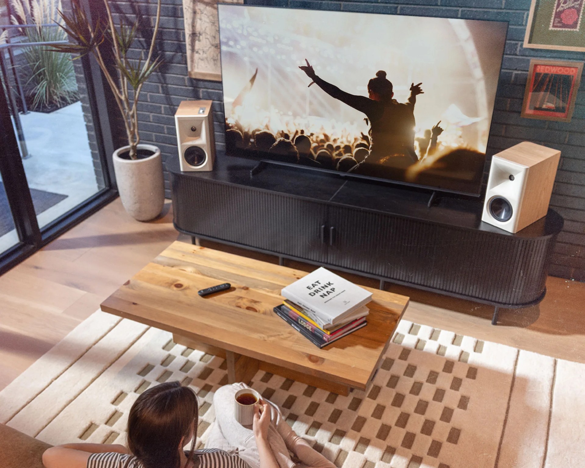Person holding a mug sitting on a beige textured rug in front of a wooden coffee table with books and a remote.