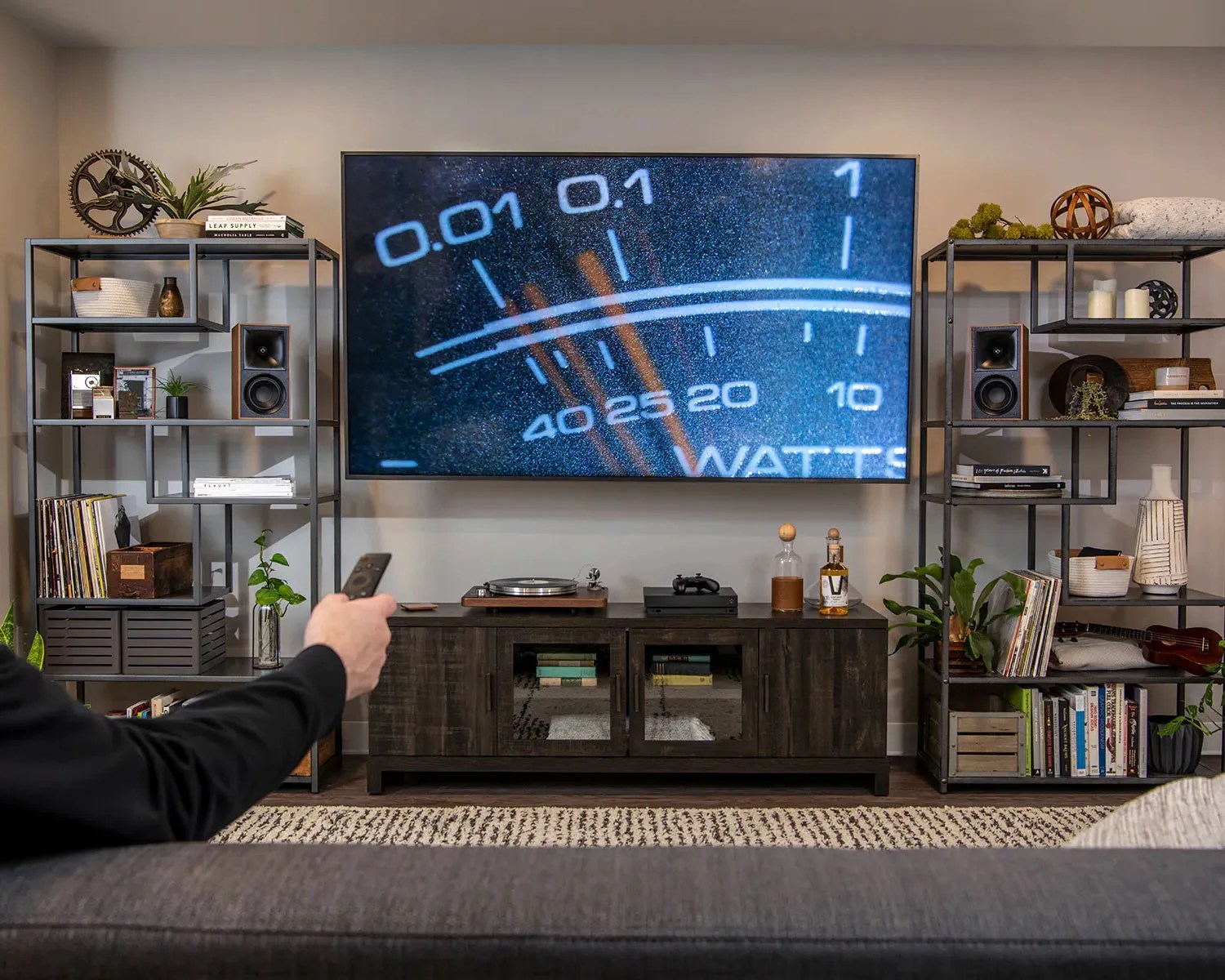Living room entertainment setup with a large wall-mounted TV displaying a close-up of a watt meter, flanked by black metal shelves with speakers and decor.