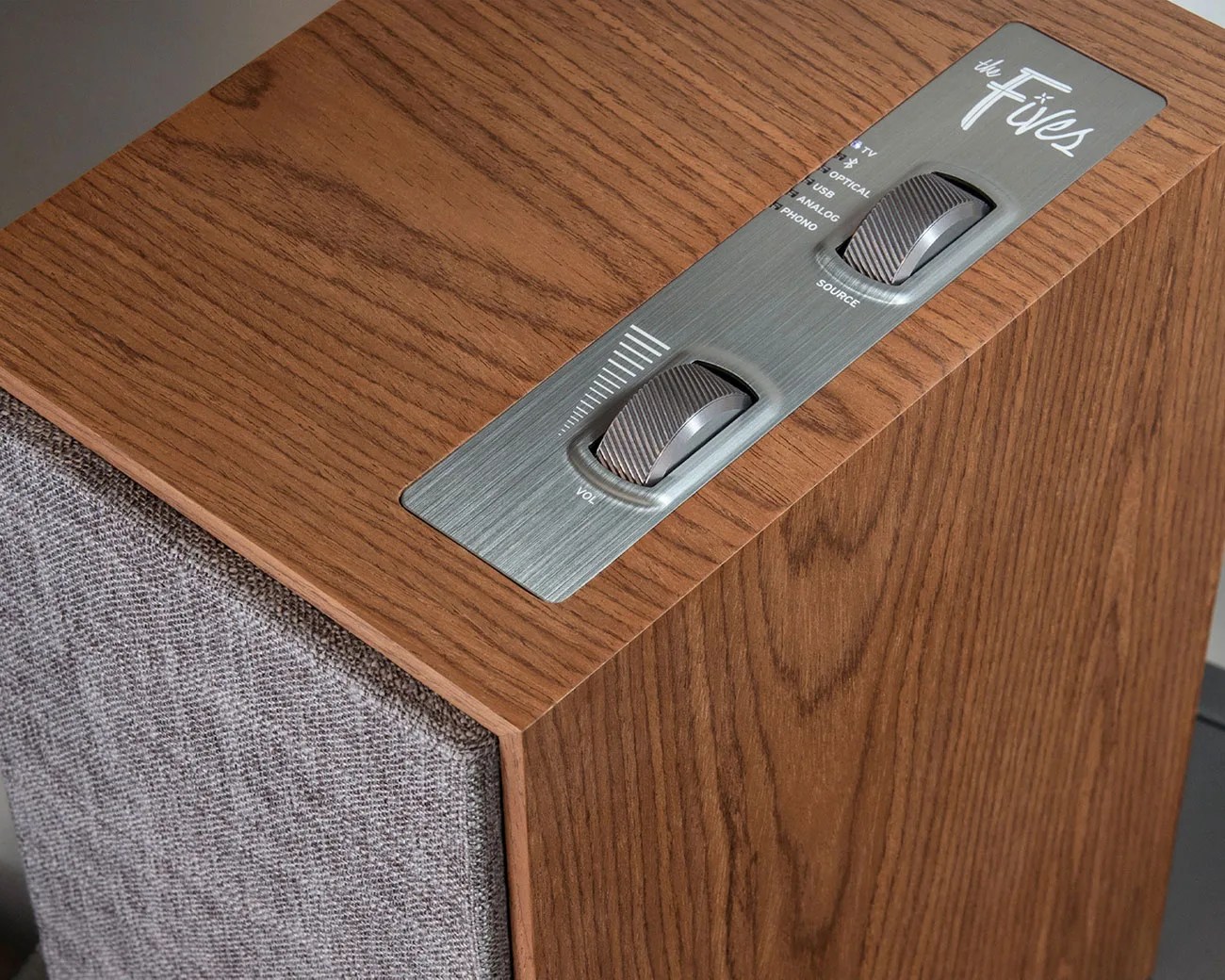 Wood-finished speaker with gray fabric front and metal control panel featuring volume and source dials.