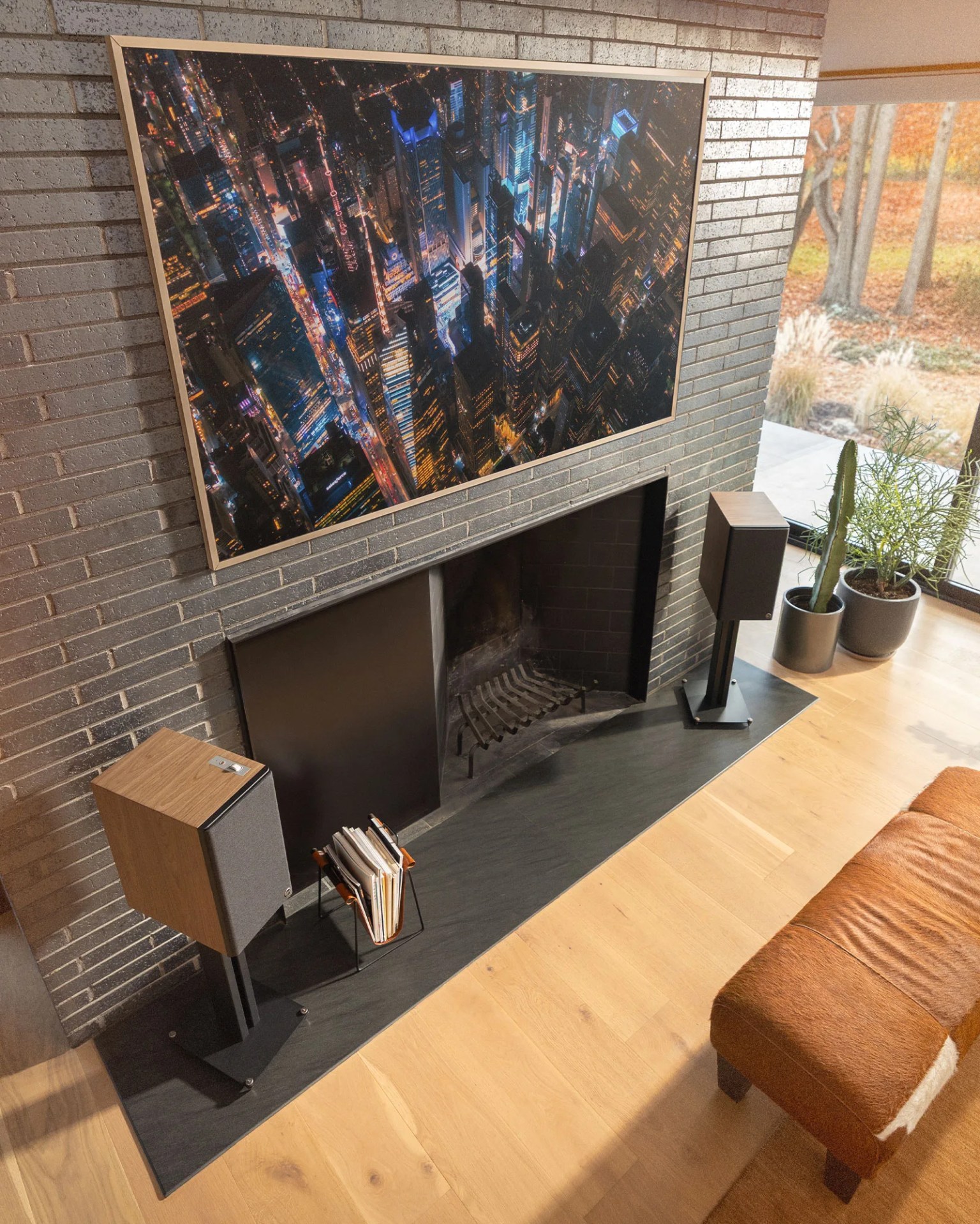 Modern living room with black brick fireplace, wooden floor, two speakers on stands, and a large cityscape photo above.