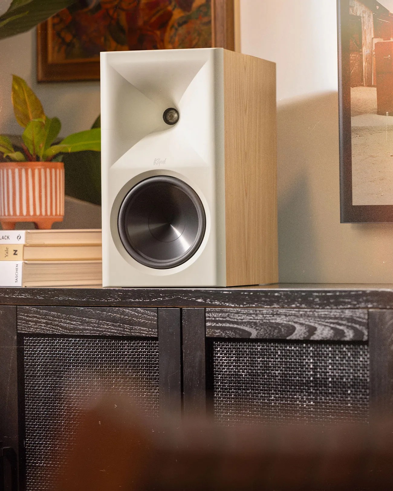 White and wood-finished Klipsch speaker on a black cabinet with books and a potted plant nearby.
