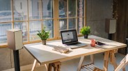 Wooden desk by a window with a laptop, two potted plants, a steaming pink mug, a tablet, and a closed book.