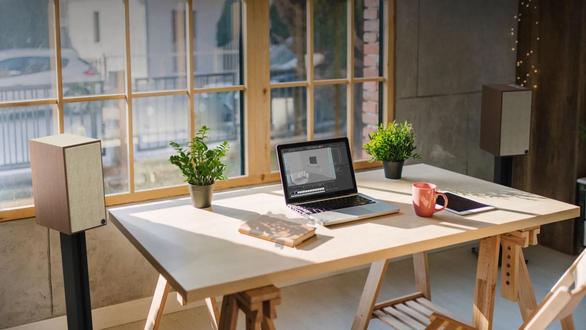 Wooden desk by a window with a laptop, two potted plants, a steaming pink mug, a tablet, and a closed book.
